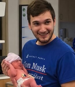 Kindyn Rheed Jinkins holding one of his newborn twin sons at the hospital, wearing a SpaceX-themed shirt — a proud father and dedicated space enthusiast, now an Ashtronaut on Space Beyond's Remembrance mission