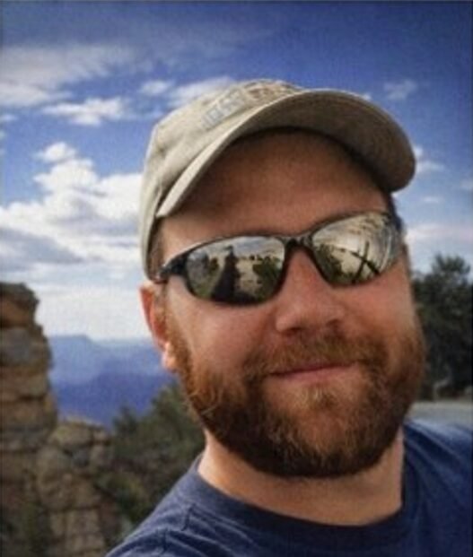 David Scott Mertins smiling in a close-up portrait, with mountains and greenery in the background.