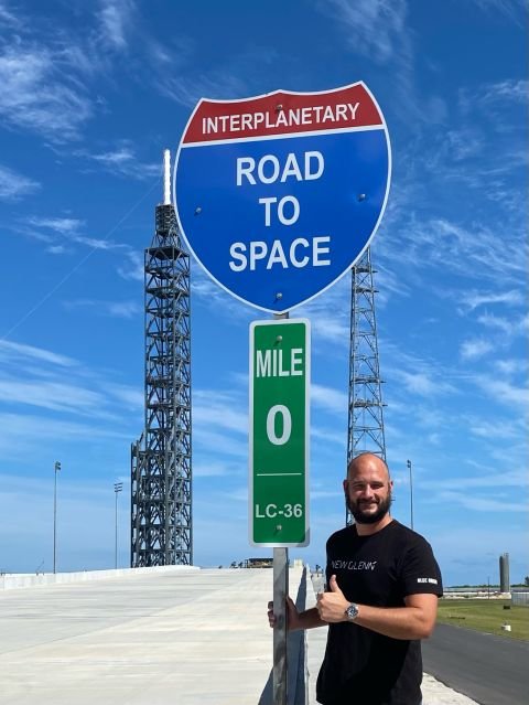 Space Beyond founder Ryan Mitchell giving a thumbs-up beside a highway-style sign reading 'Interplanetary Road to Space, Mile 0, LC-36' at Blue Origin's Launch Complex 36.