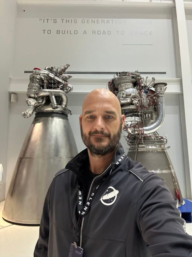 Space Beyond founder Ryan Mitchell taking a selfie in a New Glenn jacket in front of two Blue Origin rocket engine displays, beneath a wall quote about building a road to space.