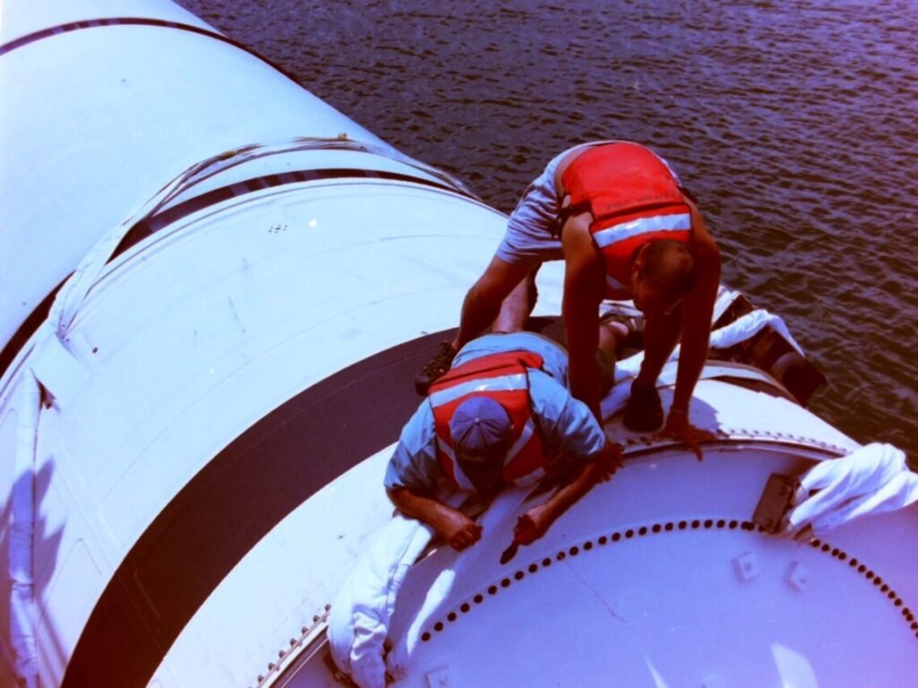 Two divers in life vests (Space Beyond founder, Ryan Mitchell, standing) working atop the forward end of a recovered Solid Rocket Booster floating in the ocean, securing it for tow back to port.