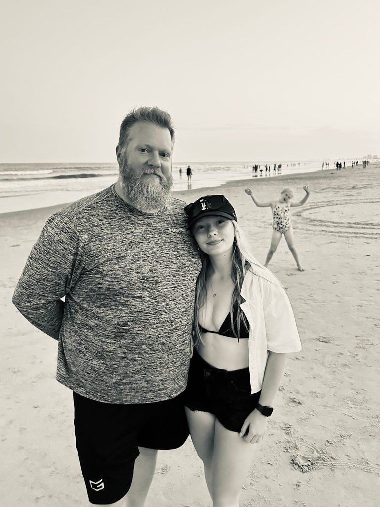 Ashtronaut Bryan J. Luther standing on the beach with his daughter, in black and white, his younger daughter playing in the background.