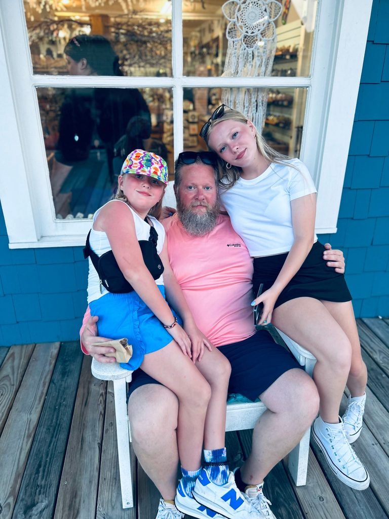 Ashtronaut Bryan J. Luther with his two daughters on a beach vacation, seated together outside a seaside shop.