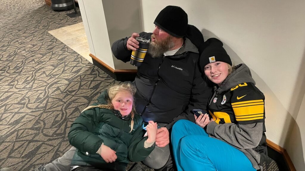 Ashtronaut Bryan J. Luther sitting on the floor with his two daughters, all three dressed for cold weather, Bryan and one daughter in Pittsburgh Steelers gear.