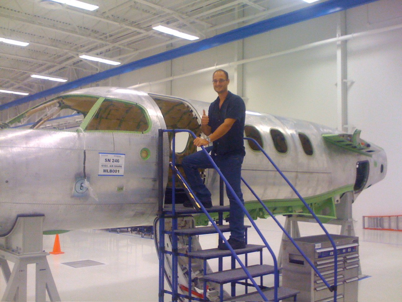 Space Beyond founder Ryan Mitchell giving a thumbs-up beside a bare, unpainted Embraer executive jet fuselage under construction on the factory floor.