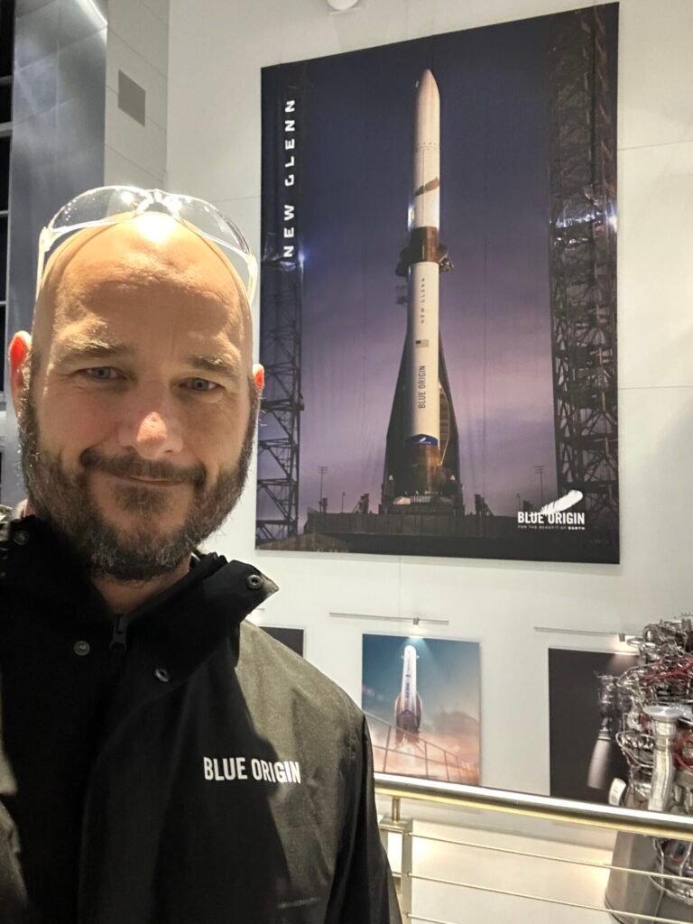 Space Beyond founder Ryan Mitchell taking a selfie in a Blue Origin jacket in front of a large New Glenn rocket poster and engine hardware on display.