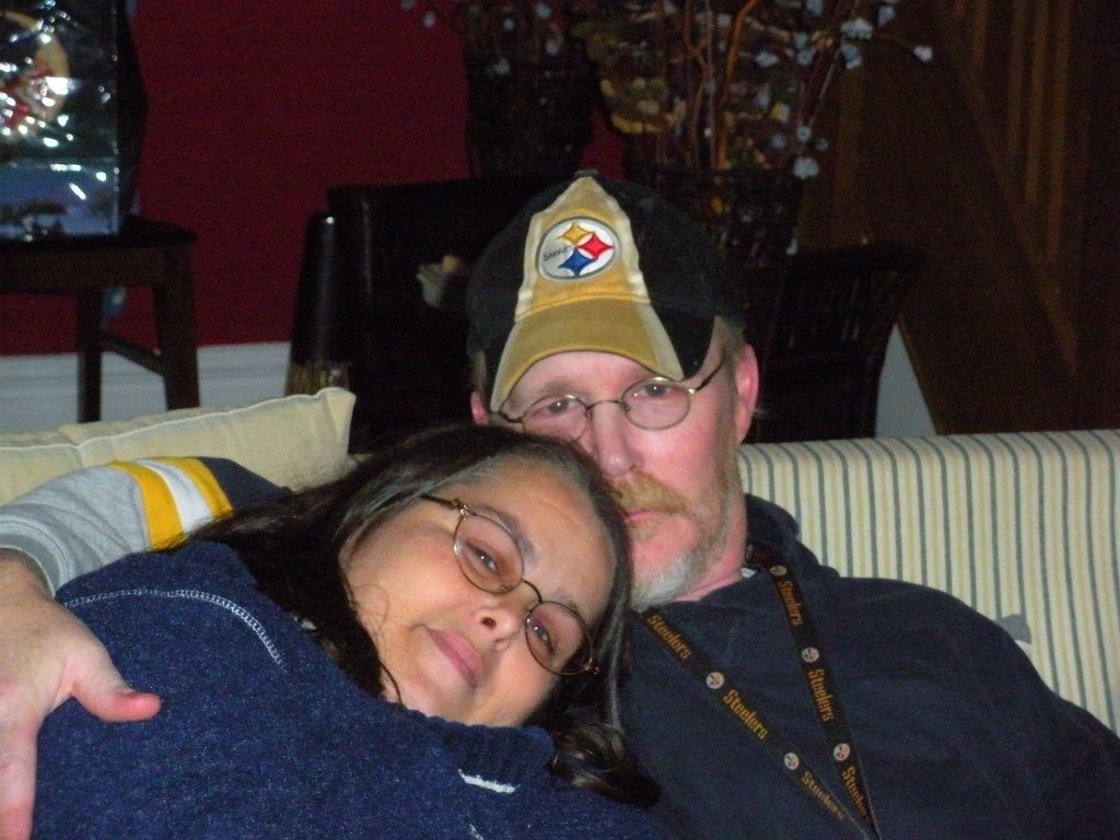 Ashtronaut Lady Gail Snow resting her head on her husband Timothy's shoulder, both smiling on a couch.