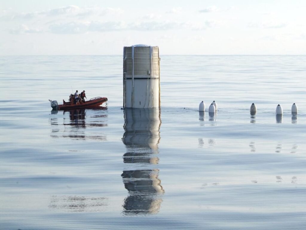 A Space Shuttle Solid Rocket Booster floating upright in the ocean after splashdown, approached by a retrieval boat and divers during recovery operations.