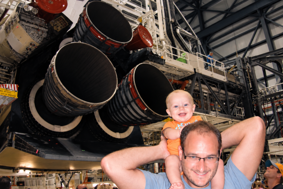 Space Beyond founder Ryan Mitchell carrying his young child on his shoulders in front of the Space Shuttle's main engines on display at the Kennedy Space Center Orbiter Processing Facility (OPF).