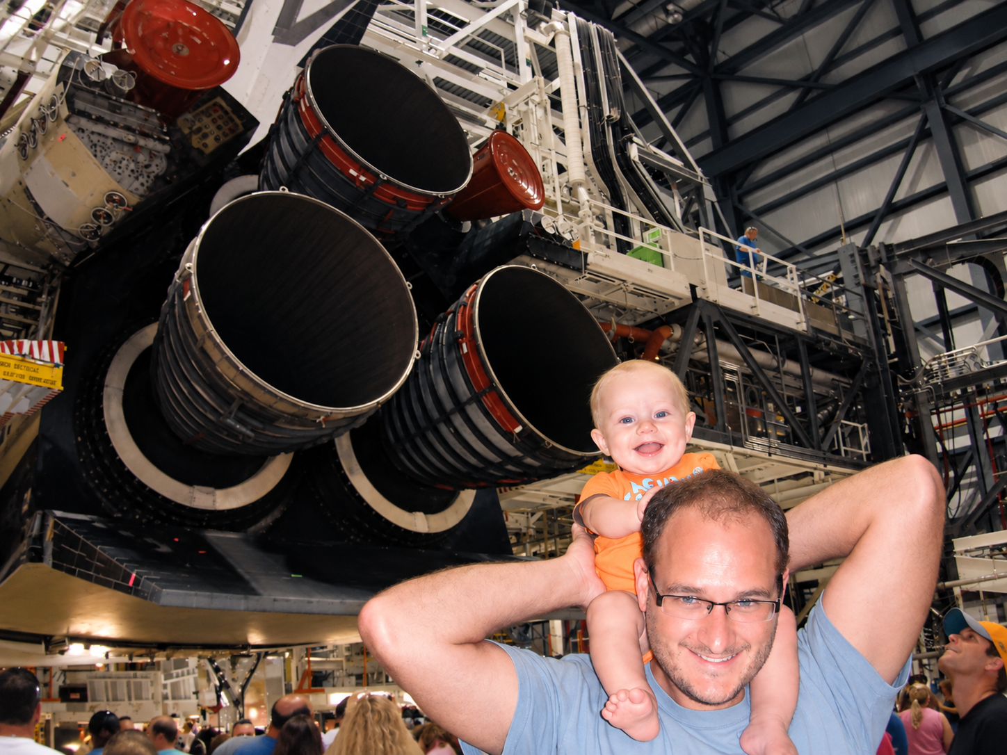 Space Beyond founder Ryan Mitchell carrying his young child on his shoulders in front of the Space Shuttle's main engines on display at the Kennedy Space Center Orbiter Processing Facility (OPF).