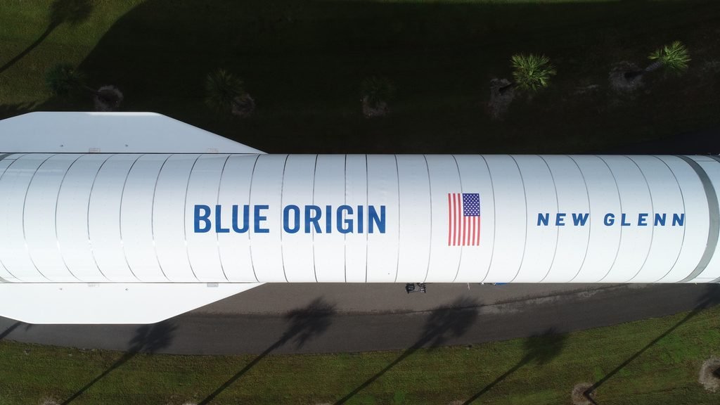 Aerial view of a full-scale New Glenn booster simulator transported outside Blue Origin's facility, painted with the Blue Origin logo, an American flag, and New Glenn lettering.