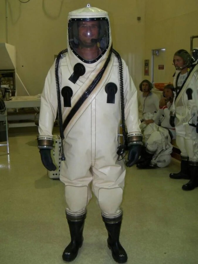 Space Beyond founder Ryan Mitchell wearing a white SCAPE suit with respirator hood and rubber boots for hazardous propellant operations at Kennedy Space Center, with two colleagues in similar suits in the background.