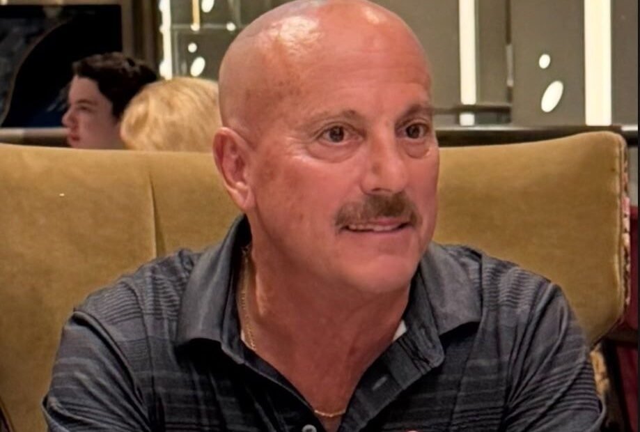 Ashtronaut Glen Scott Betterton smiling at a restaurant table, wearing a dark striped polo shirt and a thin gold chain.