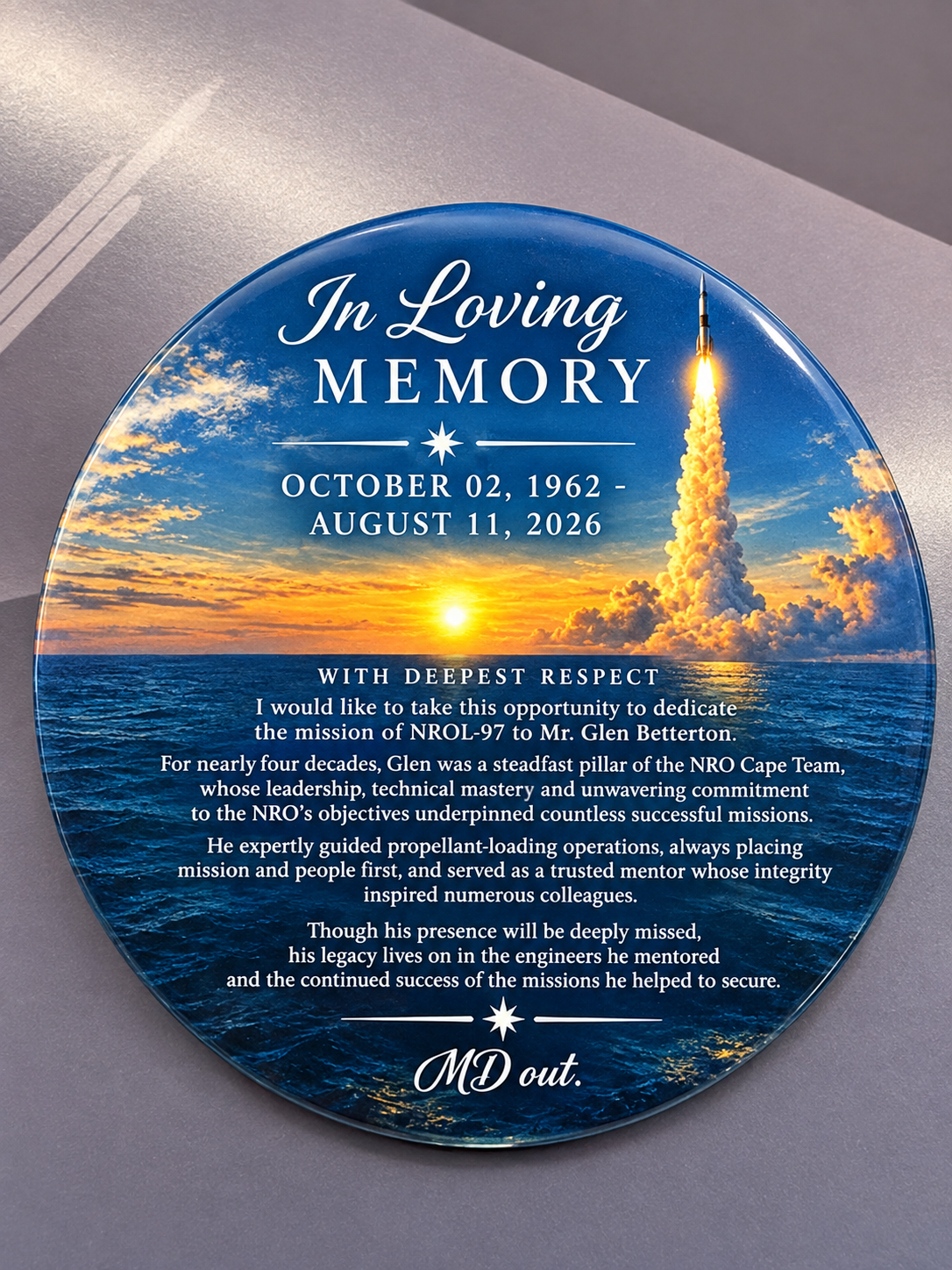 A round, glossy memorial plaque with a blue ocean and sunset background, featuring a rocket launching into the sky on the upper right. White text is arranged across the plaque. At the top it reads “In Loving MEMORY,” followed by a decorative star and the dates “OCTOBER 02, 1962 - AUGUST 11, 2026.” The center contains a memorial tribute to Glen Betterton:

“WITH DEEPEST RESPECT

I would like to take this opportunity to dedicate the mission of NROL-97 to Mr. Glen Betterton.

For nearly four decades, Glen was a steadfast pillar of the NRO Cape Team, whose leadership, technical mastery and unwavering commitment to the NRO’s objectives underpinned countless successful missions.

He expertly guided propellant-loading operations, always placing mission and people first, and served as a trusted mentor whose integrity inspired numerous colleagues.

Though his presence will be deeply missed, his legacy lives on in the engineers he mentored and the continued success of the missions he helped to secure.”

A decorative star appears beneath the tribute, followed by the signature “MD out.” The plaque is photographed resting on a gray surface.